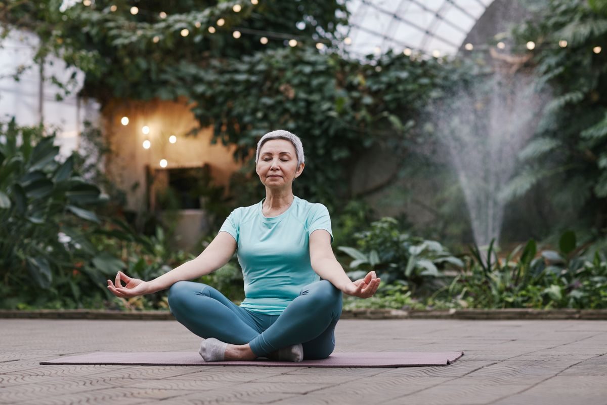 A smiling senior woman sitting cross-legged on a yoga mat in a bright greenhouse, hands resting on her knees